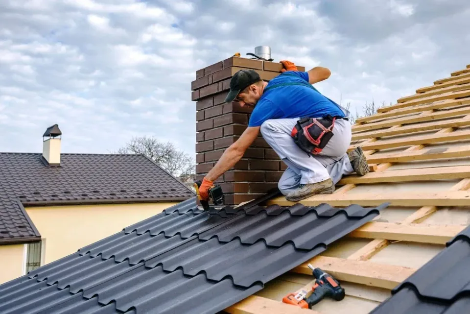 Roofer in blue shirt installing dark metal roof shingles on a pitched roof, with brick chimney and wooden framing visible.